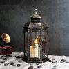 A vintage Nordic iron candle lantern with glass panes sits on a gray cloth surrounded by smooth stones.