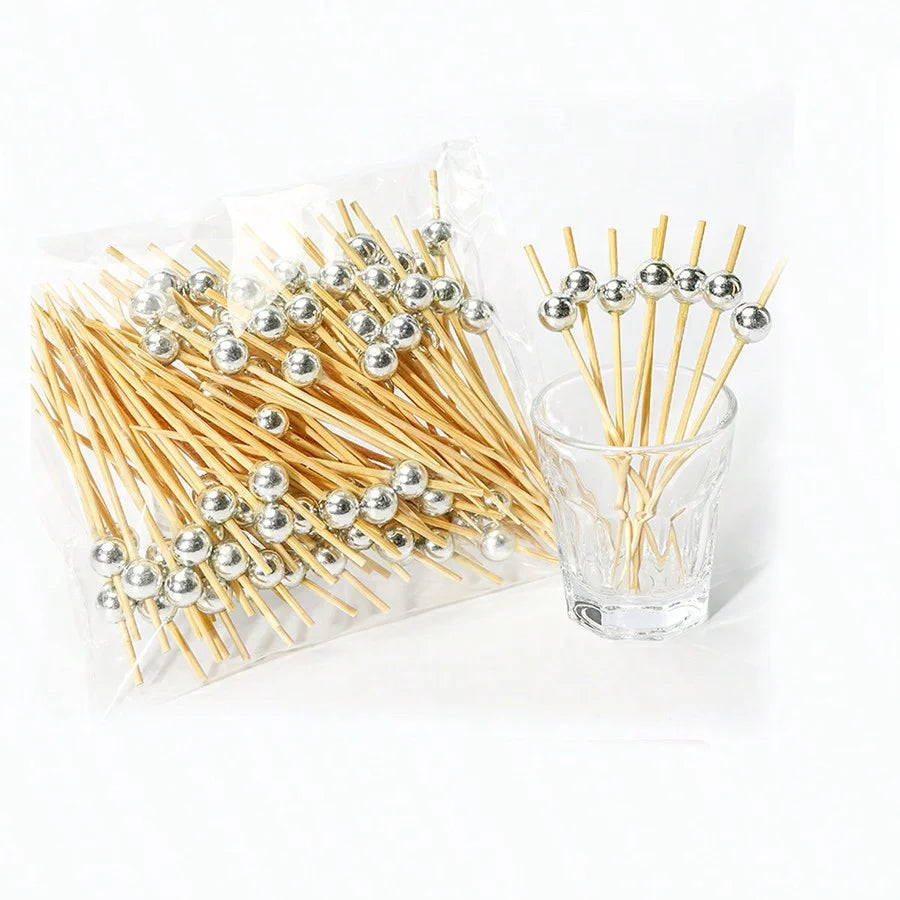 A clear plastic bag filled with bamboo fruit skewers featuring silver ball tops, arranged next to a glass.