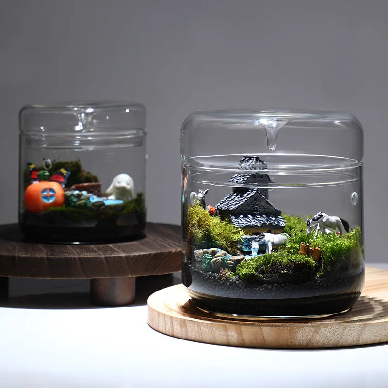 A clear glass vase filled with moss, rocks, and a miniature house structure for decoration.