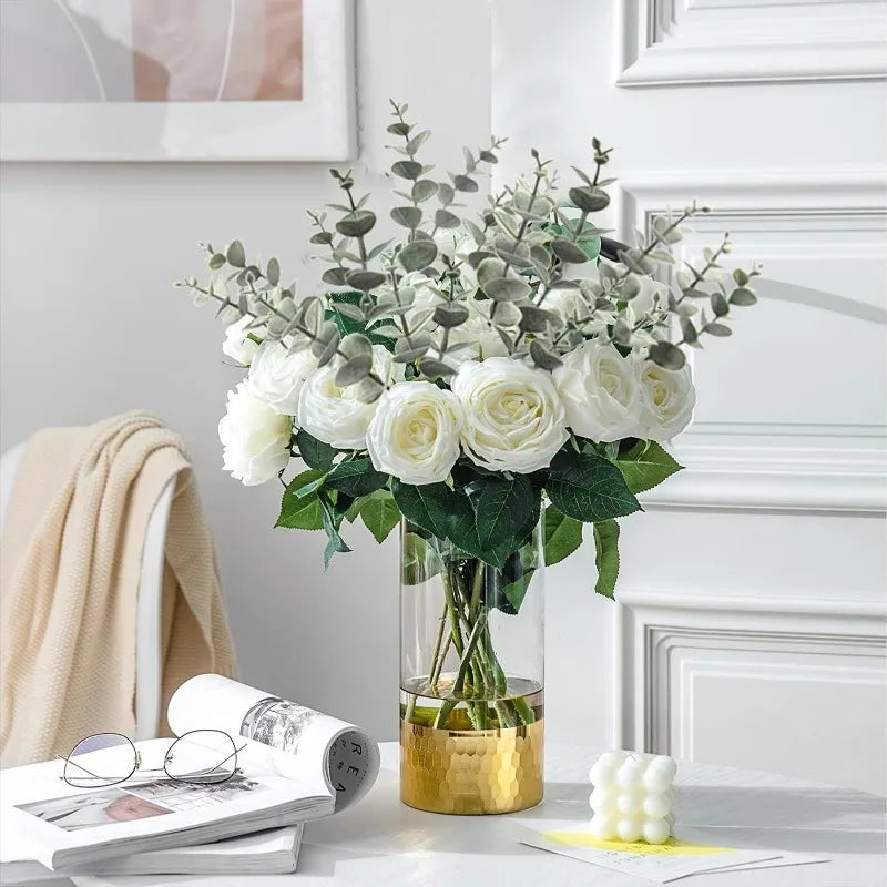 A bouquet of white roses and silvery-green eucalyptus stems in a glass vase on a white surface.