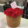 A bouquet of deep red artificial foam roses in a brown paper container with a handwritten tag.
