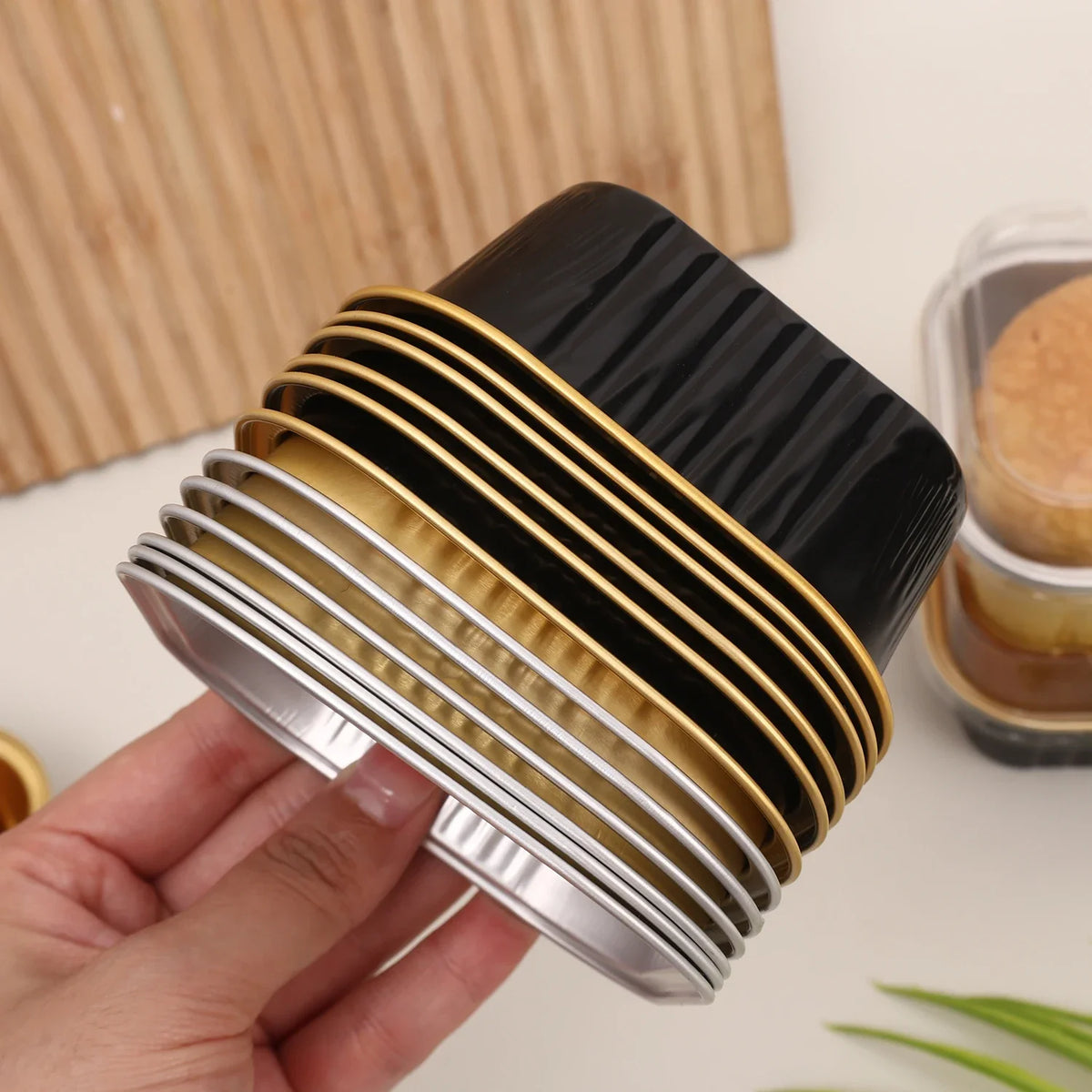 A hand holds a stack of square aluminum foil baking boxes with transparent lids in silver and gold tones.