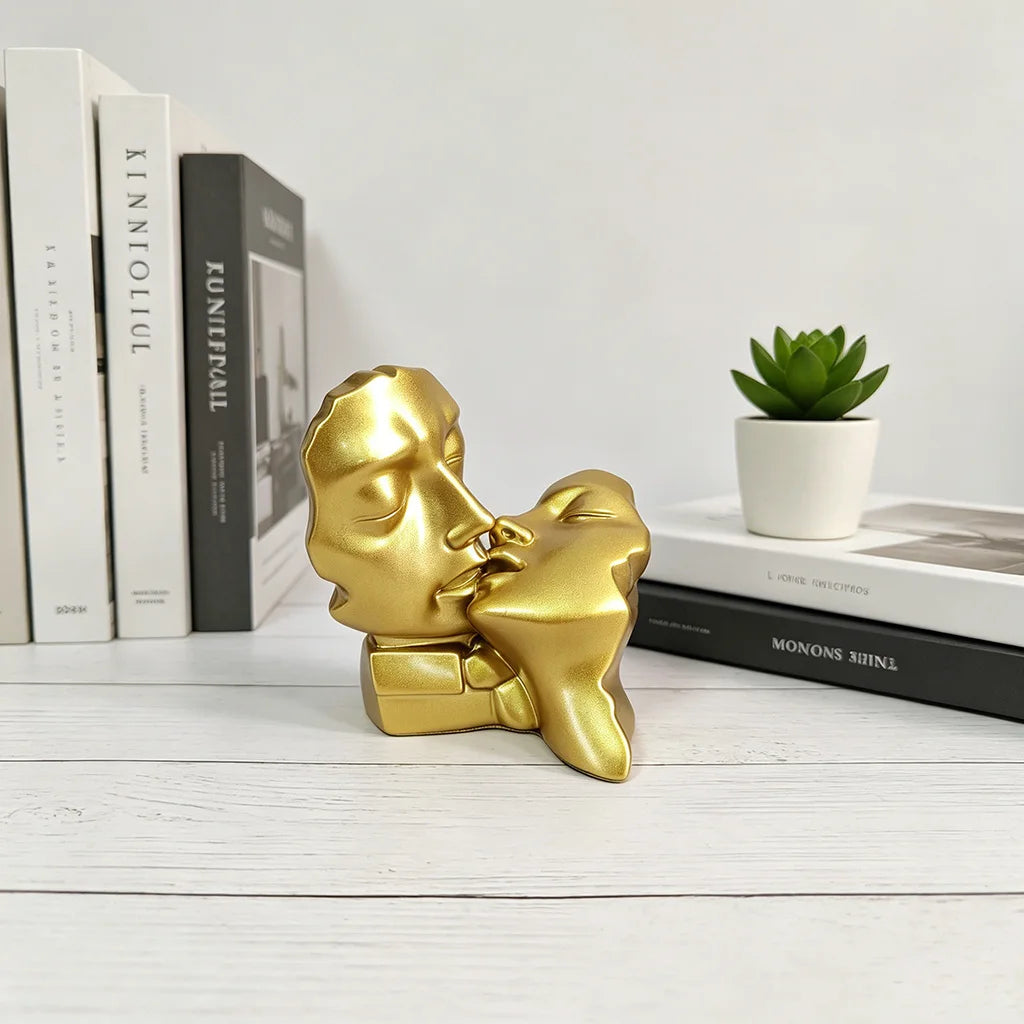 A golden resin sculpture of a kissing couple sits on a white wooden surface next to books and a potted plant.