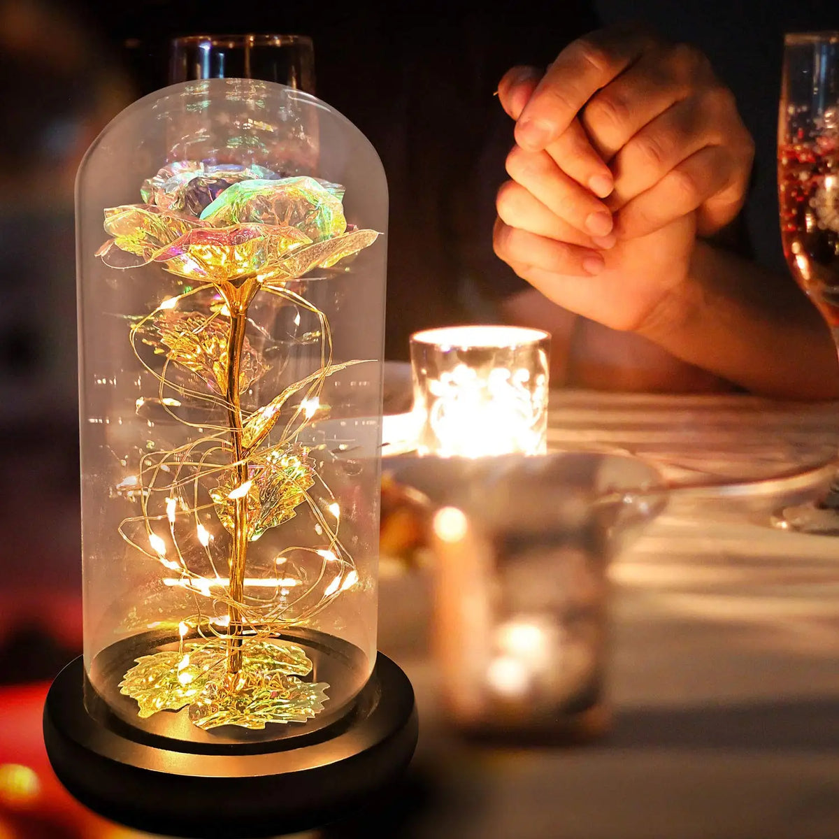 Une rose de verre éternelle avec un tige doré et des lumières LED est exposée sur une surface sombre à côté d'une bougie.