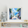 A framed painting depicting a sunny coastal street with white buildings, lush greenery, and blue sea views.