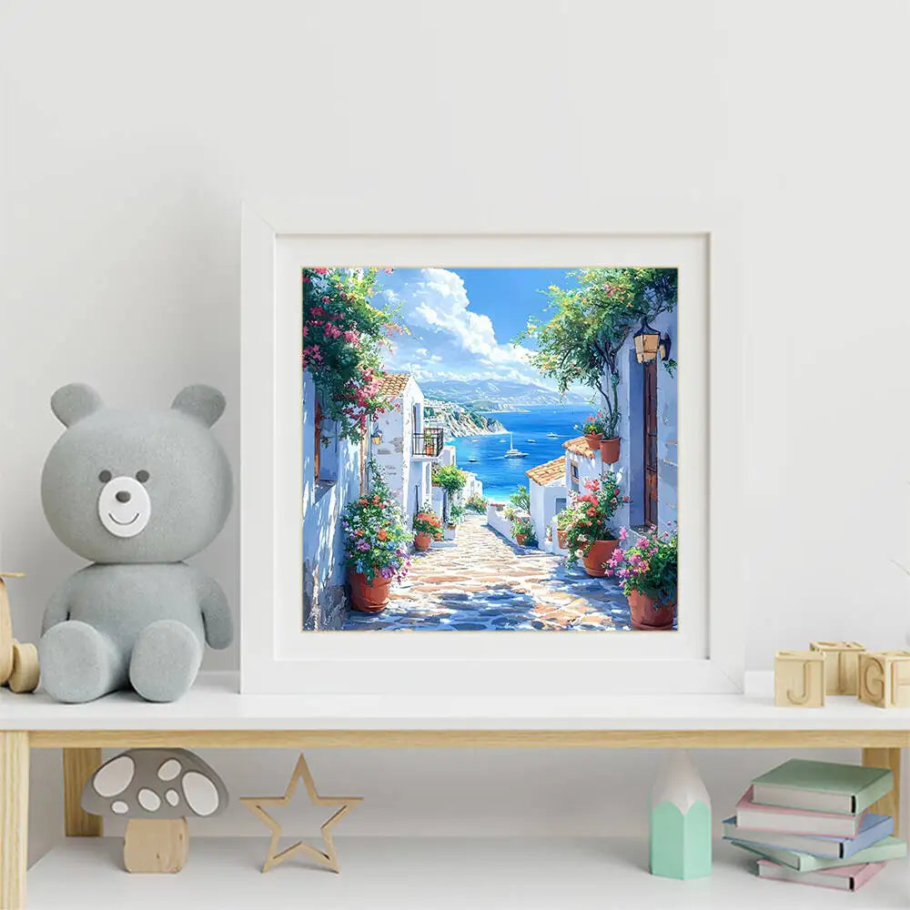 A framed painting depicting a sunny coastal street with white buildings, lush greenery, and blue sea views.