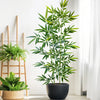 A tall artificial bamboo branch with green leaves displayed in a black pot against a white background.