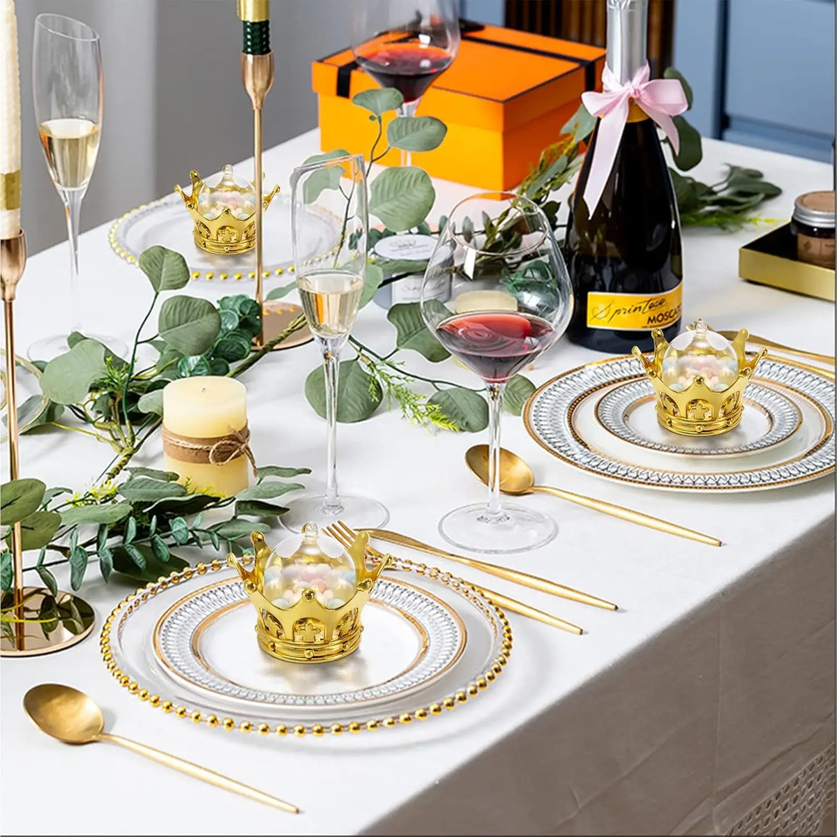 A table setting featuring gold crown candy boxes, silver plates, and glassware for a formal event.