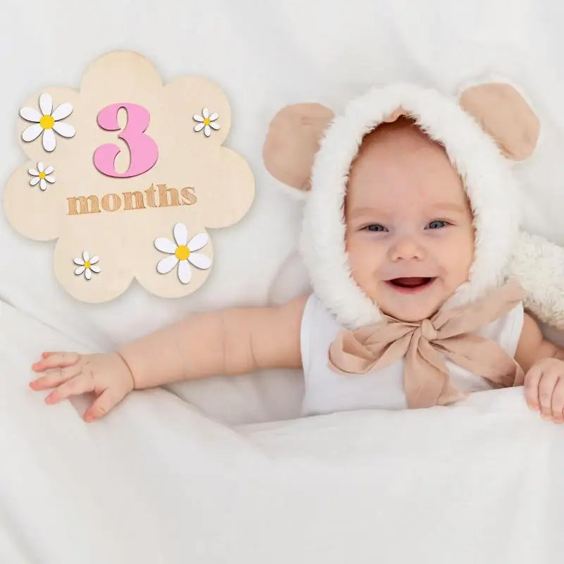 A smiling baby wearing a fluffy bear headband lying on white fabric next to a wooden milestone card marking three months.