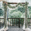 An archway decorated with artificial eucalyptus leaf garlands and white fabric in an outdoor setting.