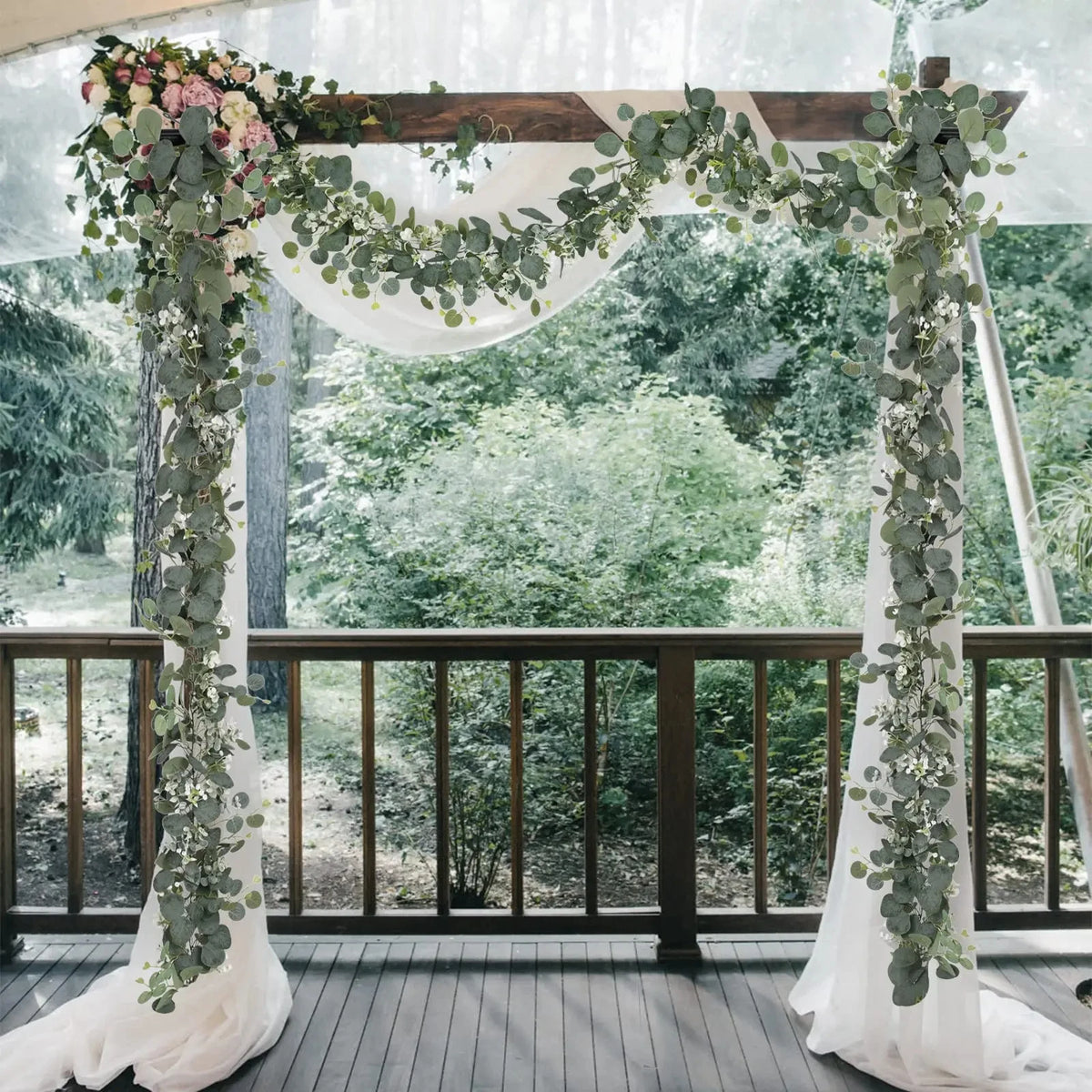 An archway decorated with artificial eucalyptus leaf garlands and white fabric in an outdoor setting.