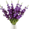 Un bouquet de fleurs artificielles de delphinium violet avec du feuillage vert dans un pot blanc.