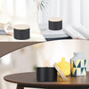A set of black metal candle tins with lids displayed on a wooden table next to a yellow glass and a book.