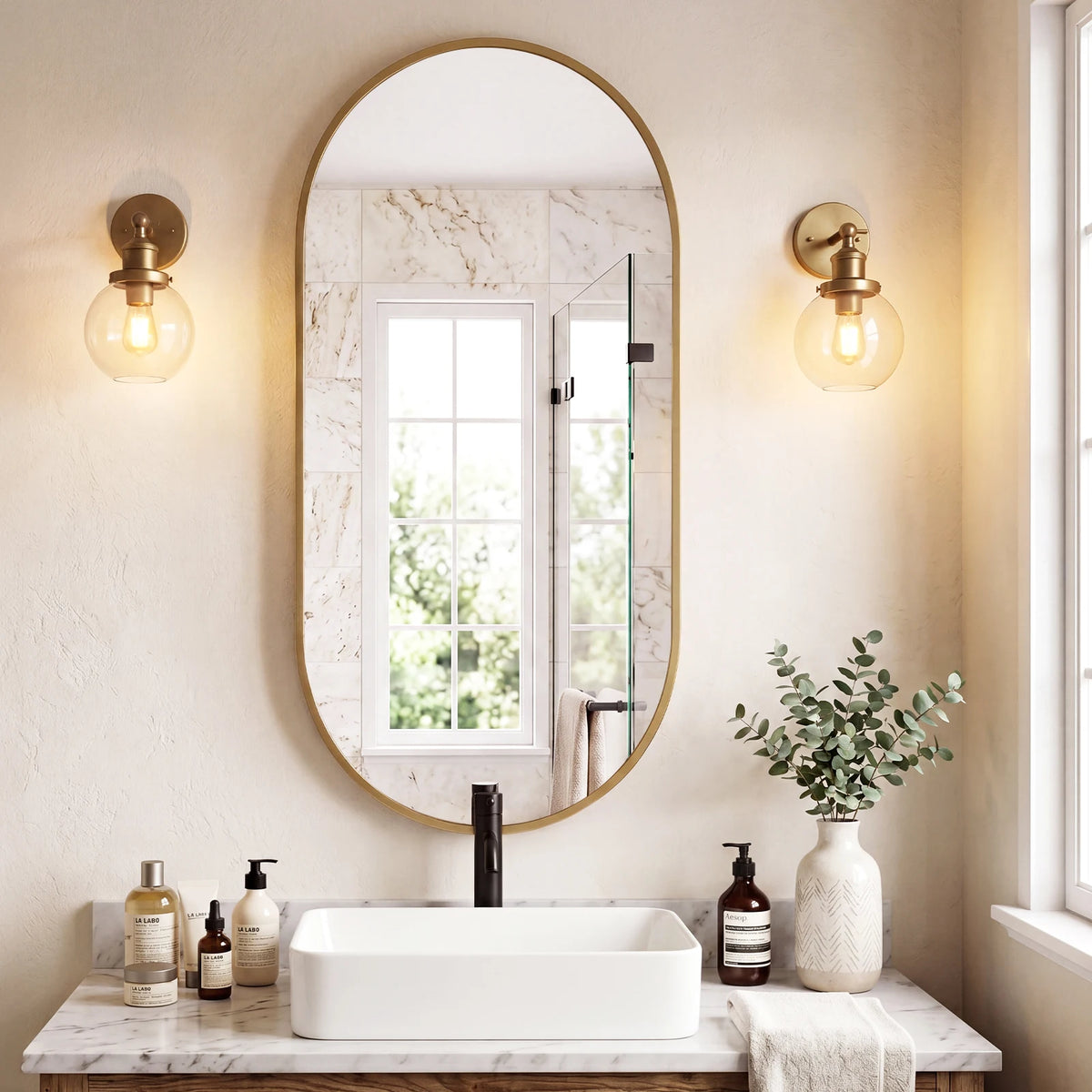 A bathroom vanity featuring an oval mirror with a gold frame, a white vessel sink, and natural light fixtures.