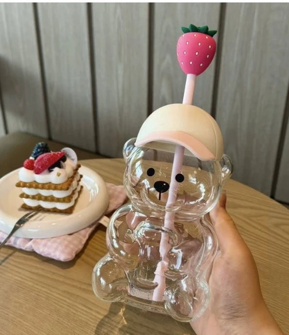 A clear glass coffee cup with a strawberry-shaped straw and a bear-shaped glass holder is held in a hand.