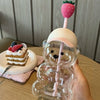 A clear glass coffee cup with a strawberry-shaped straw and a bear-shaped grip is held in a hand.