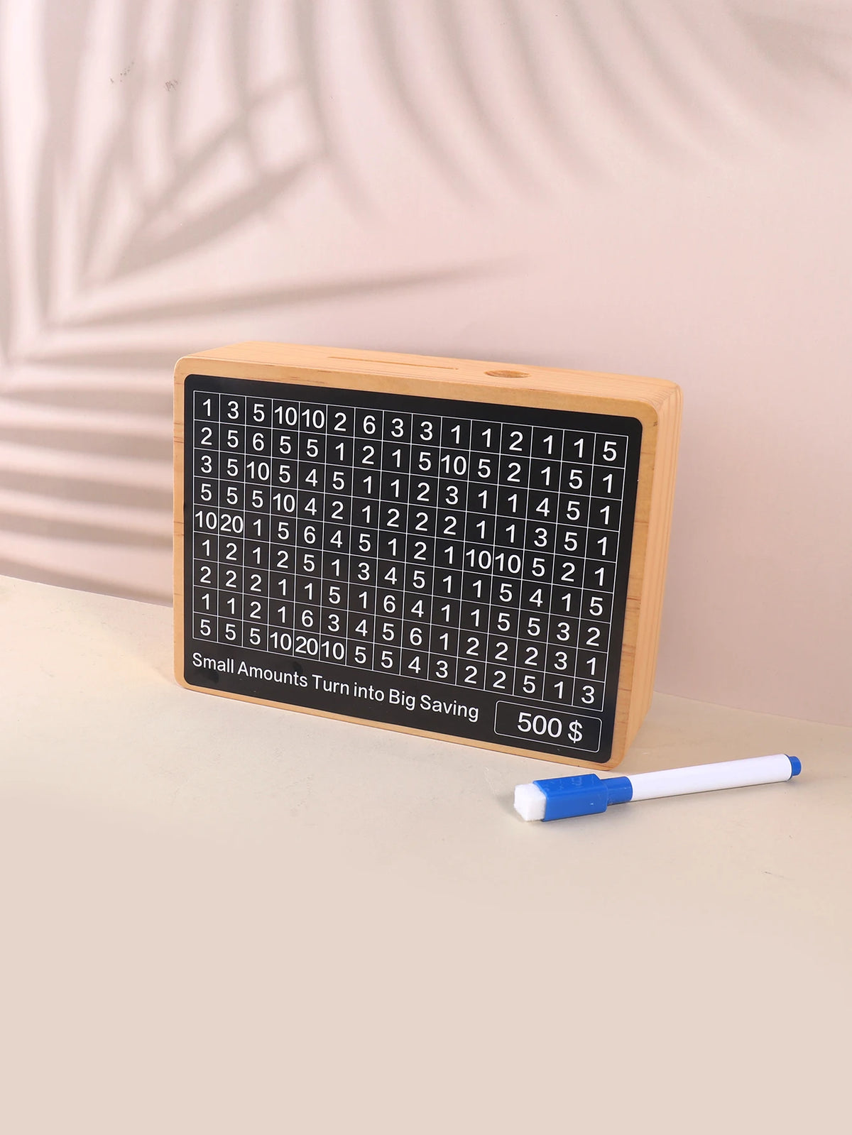 A wooden savings box with a grid of numbers is shown next to a blue pen.