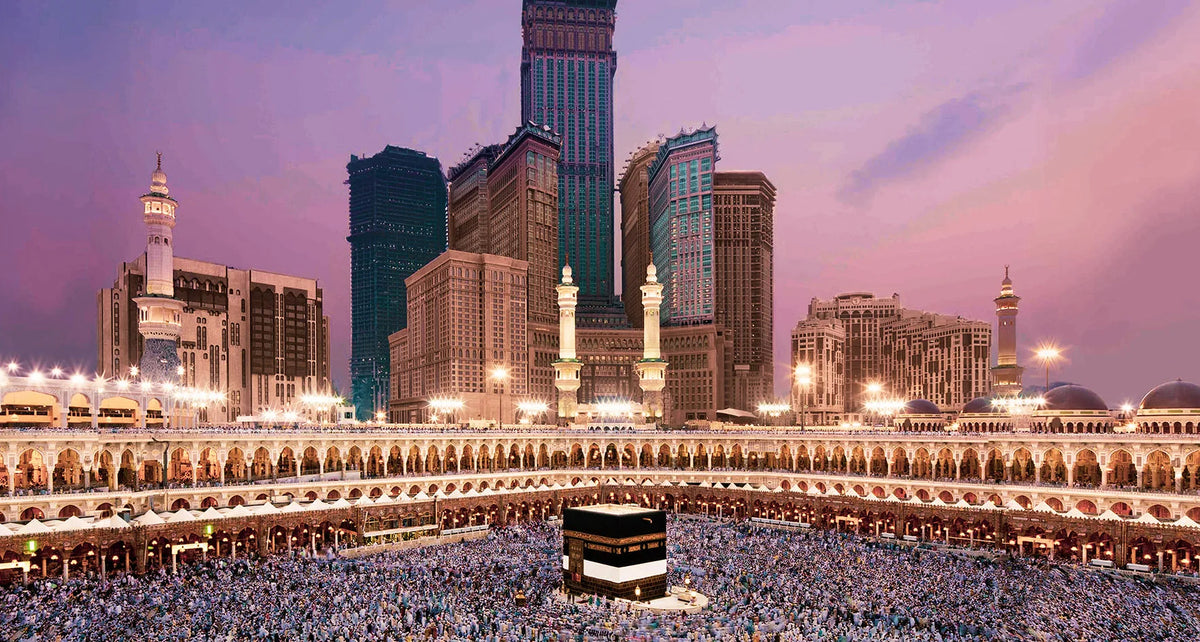 A panoramic view of a large mosque complex with modern skyscrapers in the background, featuring a large crowd in the foregrou