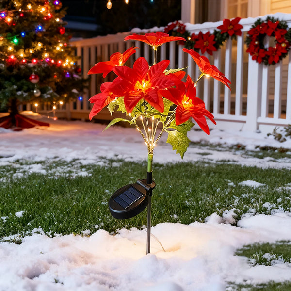 A solar-powered poinsettia garden light with bright red flowers illuminates a snowy lawn in front of a white fence.