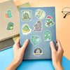 A hand holds a sheet of Kawaii Green Dinosaur PVC stickers for scrapbooking and stationery on a blue surface.