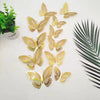 A set of twelve gold glitter butterfly wall stickers scattered on a white surface next to a green plant and a palm leaf.