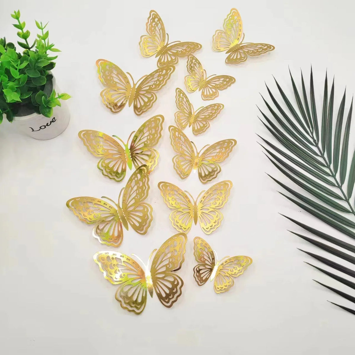 A set of twelve gold glitter butterfly wall stickers scattered on a white surface next to a green plant and a palm leaf.