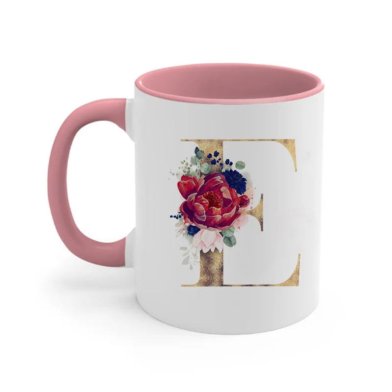 A pink ceramic coffee mug featuring a large gold letter E decorated with floral elements.