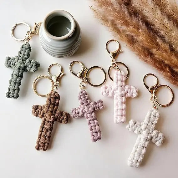 A set of bohemian woven cross keychains in various shades of green, beige, and pink, displayed next to a ceramic cup.