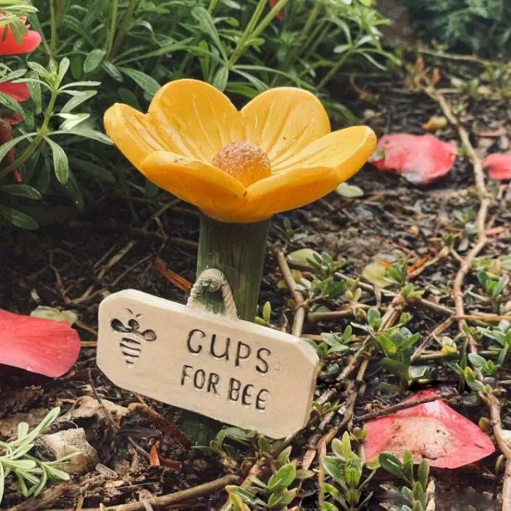 A yellow flower with a golden center is growing in the dirt, accompanied by a wooden tag labeled Gups for Bee.