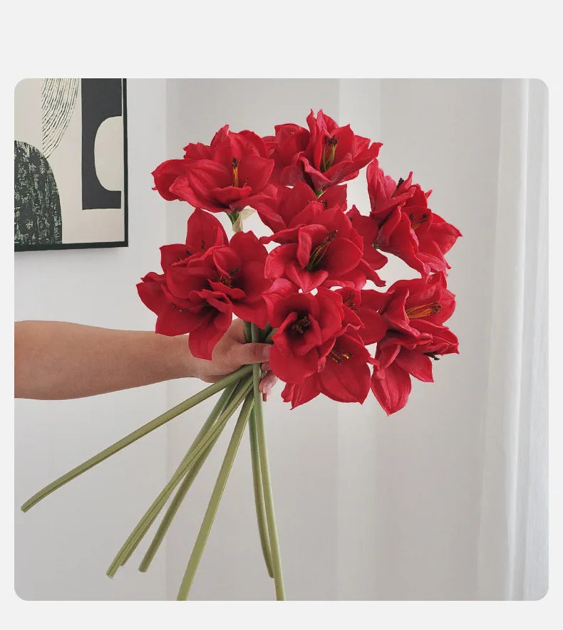 A hand holds a bouquet of vibrant red artificial clivia amaryllis flowers against a white background.