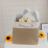 A beige fabric storage cube with a light blue stuffed animal inside, next to a yellow sunflower.