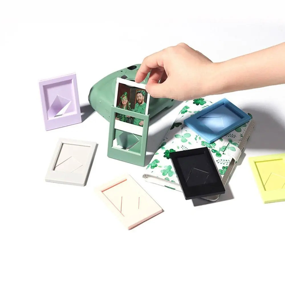 A hand is holding a mini film frame displaying instant photos, with various colored frames scattered on a white surface.