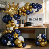 A balloon arch display featuring navy blue, white, and gold balloons arranged in a crown shape with academic props.