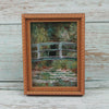 A framed painting featuring a black cat sitting on a wooden bridge in a natural, mossy landscape.