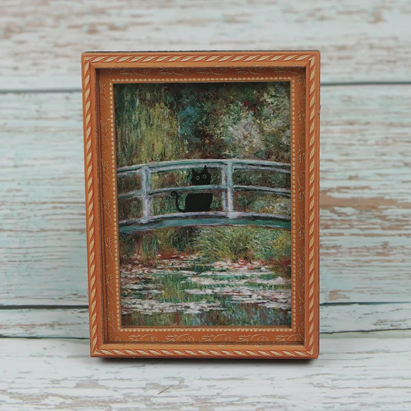 A framed painting featuring a black cat sitting on a wooden bridge in a natural, mossy landscape.