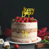 A round cream-colored birthday cake decorated with fresh strawberries and a gold Happy Birthday topper.