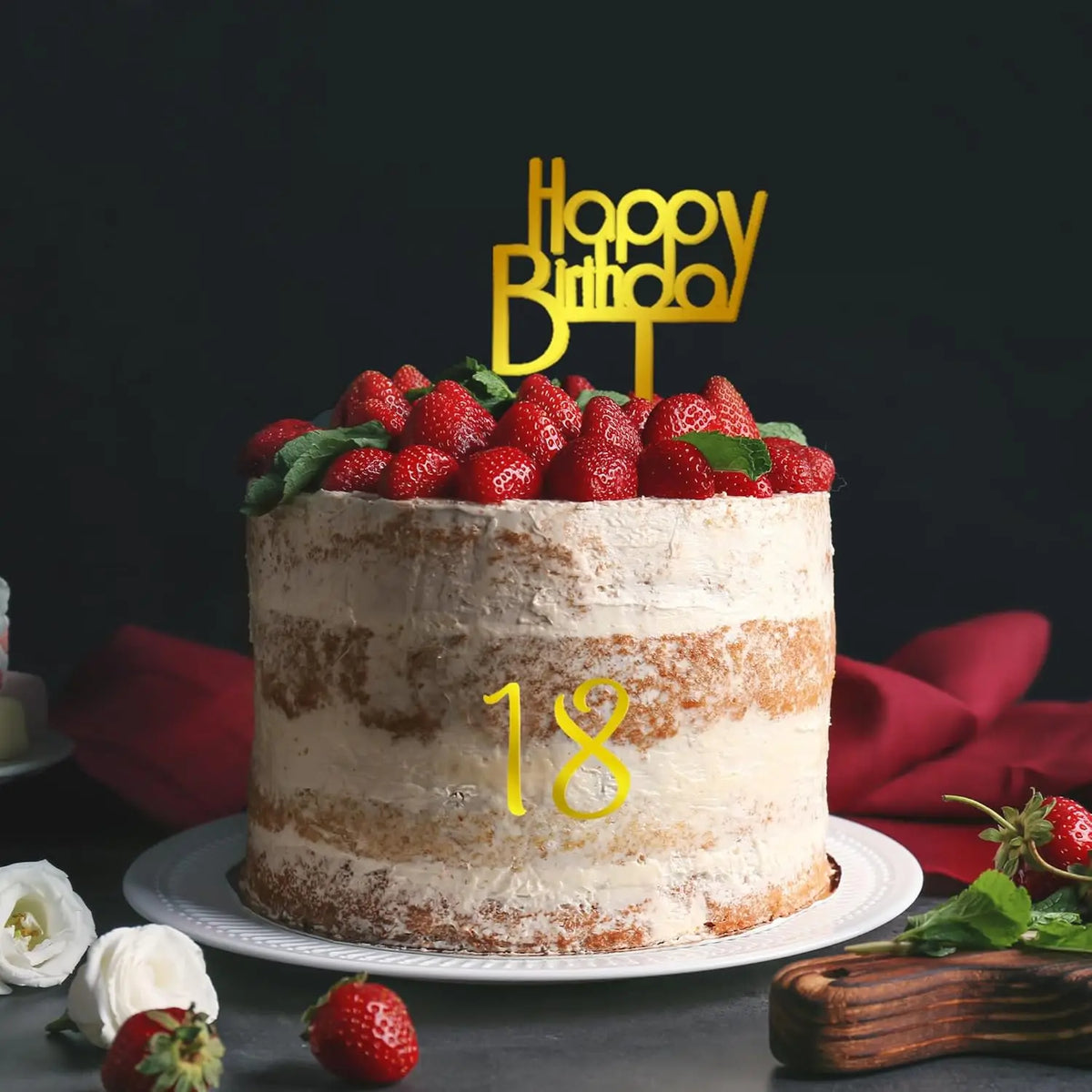 A round cream-colored birthday cake decorated with fresh strawberries and a gold Happy Birthday topper.
