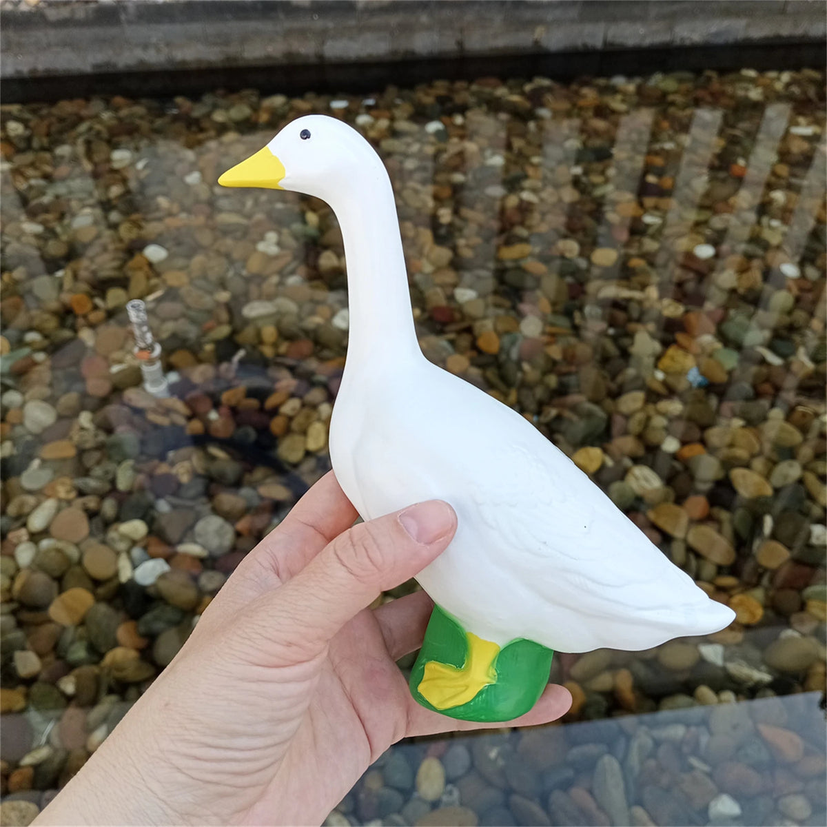 A white garden statue of a goose with yellow accents is held above a bed of small stones.