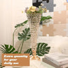 Goldener Kristallvase mit lila und gelben Blumen auf weißer Fläche neben grünen tropischen Blättern.