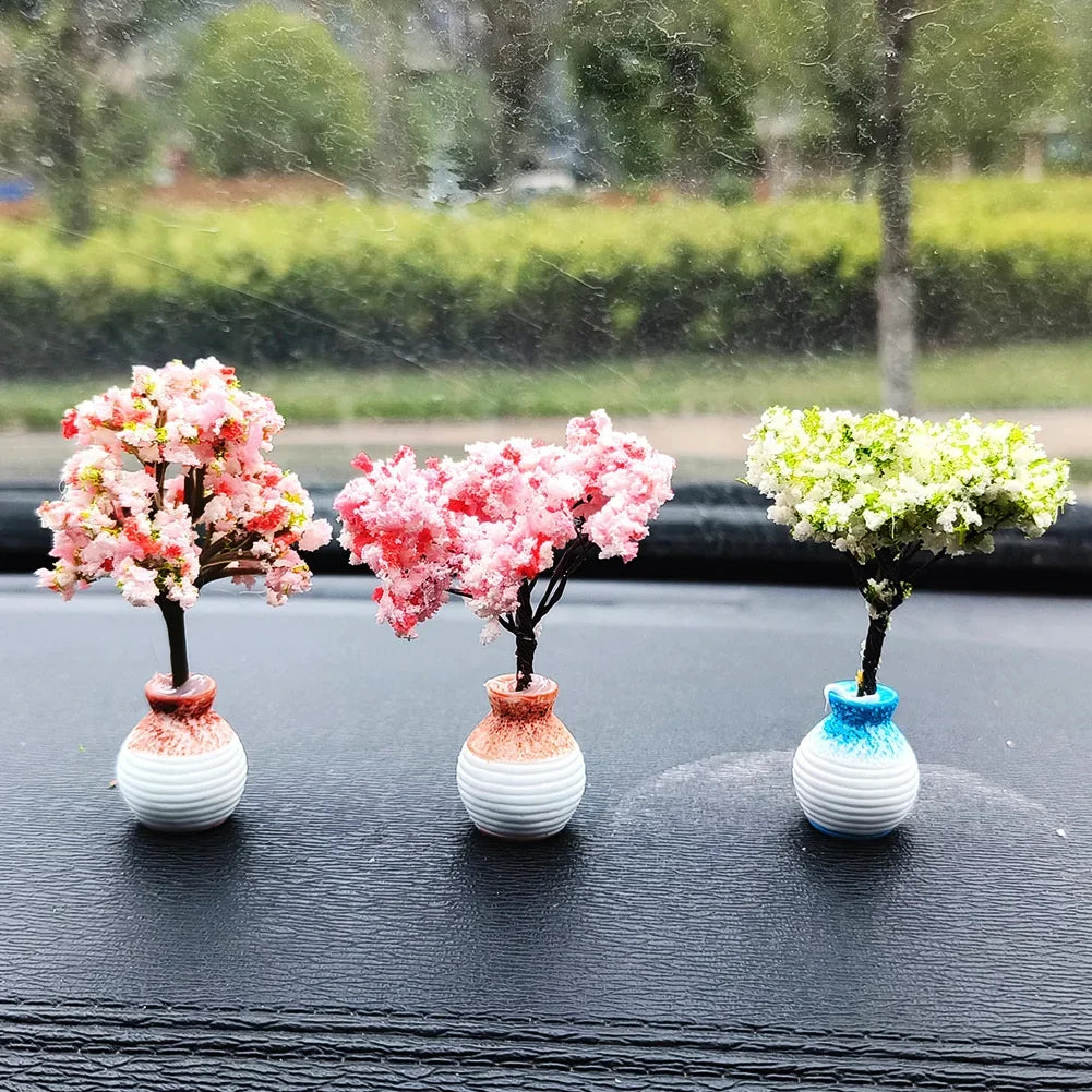 Three miniature tree ornaments with pink and white flowers displayed on a black car dashboard.