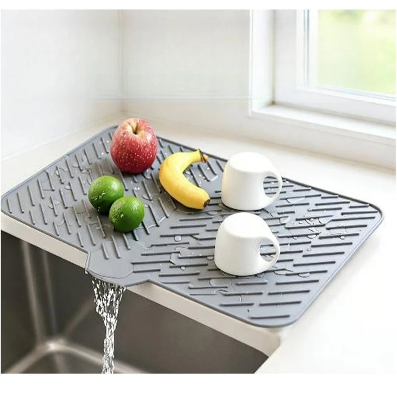 A silicone drain mat in gray with textured ridges sits on a kitchen sink, holding fruit and a white ceramic pitcher.