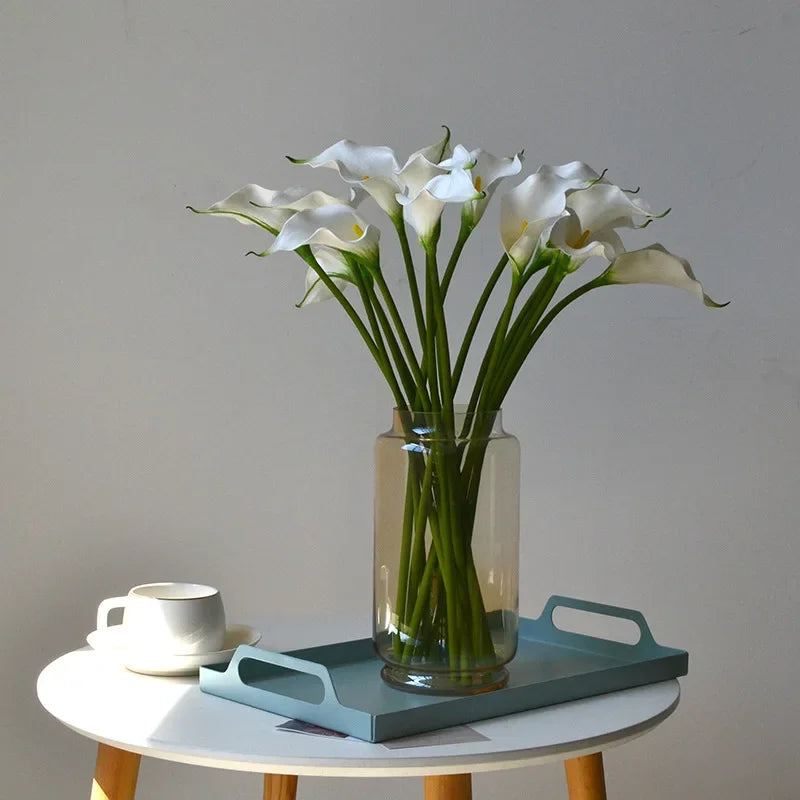 A bouquet of white artificial calla lily stems in a clear glass vase on a light-colored table.