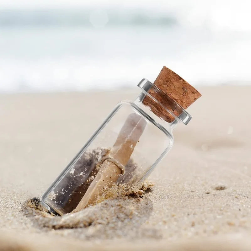 A glass vial with a cork stopper and some light-colored powder is pressed into the sand on a beach.