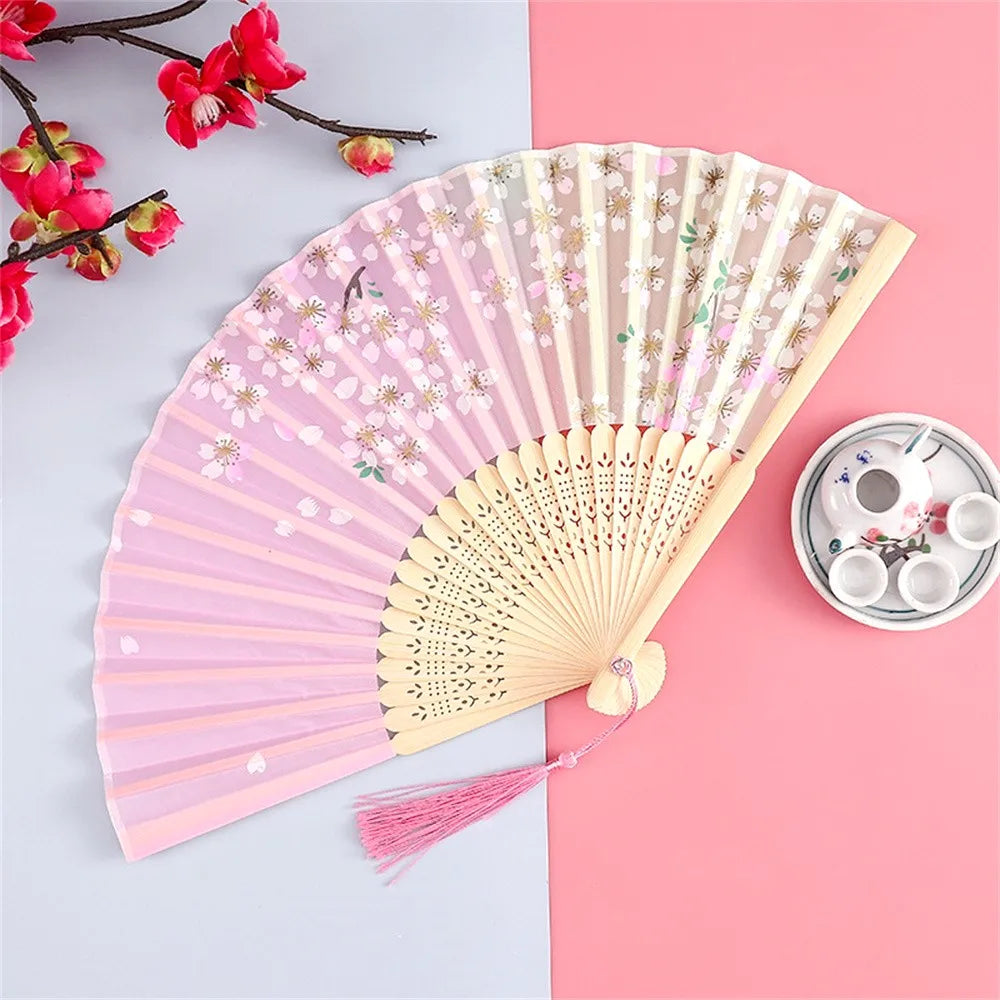 A pink and gold vintage silk folding hand fan is displayed on a light background next to a small white teacup set.