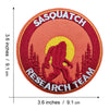 An embroidered patch featuring a red Bigfoot silhouette against a yellow sun, with the text SASQUATCH RESEARCH TEAM.
