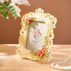 A vintage gold-framed resin photo frame decorated with roses and greenery, resting on a wooden surface.