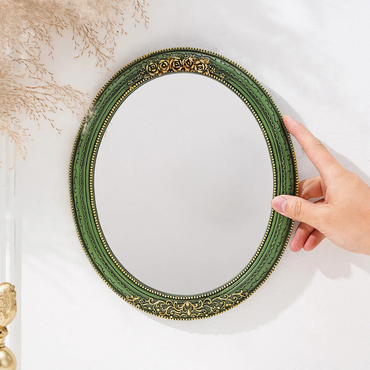 A close-up of a vintage green oval wall mirror with an intricate gold lace design, being held by a hand.