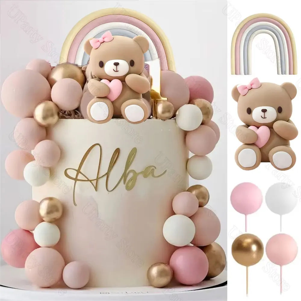 A cake decorated with pink and gold ball decorations and a teddy bear topper, set against a white background.