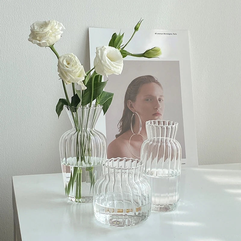 A clear glass vase holds white roses and is displayed next to smaller glass vessels.