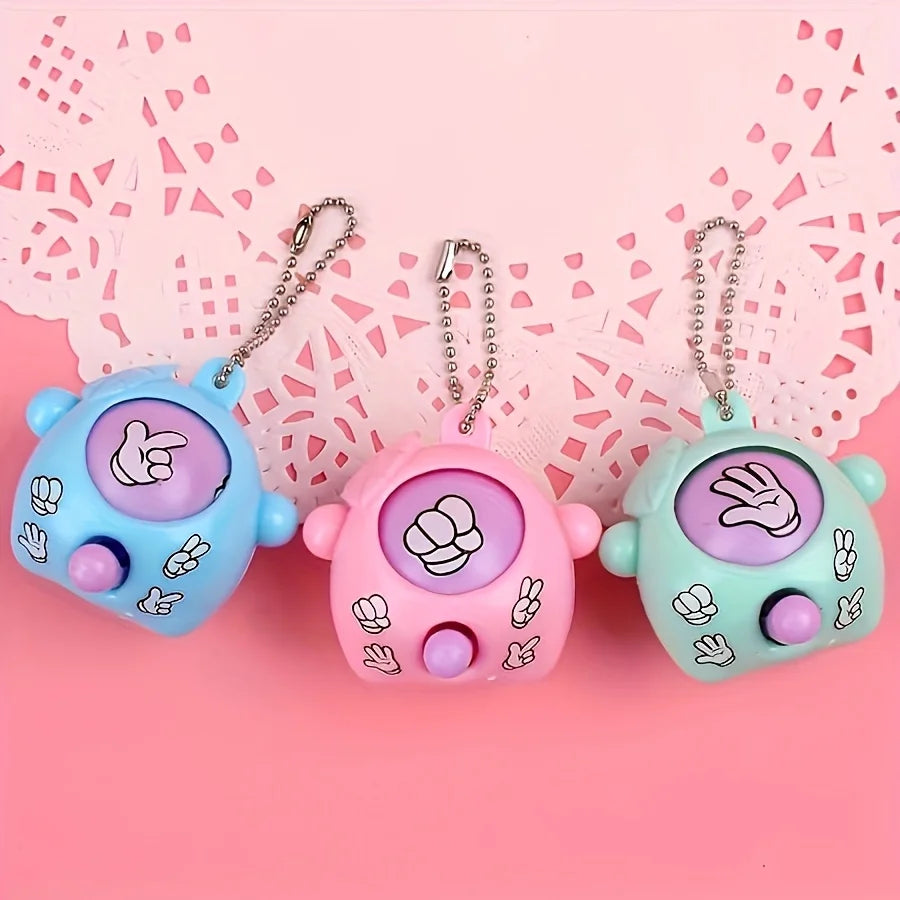 Three colorful mini rock paper scissors keychains with hand gestures hanging against a pink lace background